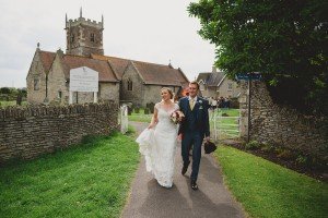 A bride and groom joyfully walk down a path outside a stone church, captured by talented wedding photographers Somerset. The bride holds a bouquet in her white dress, while the groom, in a blue suit with a top hat, beams happily. A small group gathers near the church entrance amidst lush greenery.