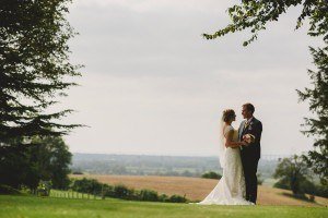 A bride and groom stand closely together in a landscape setting captured beautifully by Somerset wedding photographers. They are on a grassy area with trees framing the image, while fields stretch into the distance under a cloudy sky. The bride holds a bouquet, adding charm to this elegant moment.