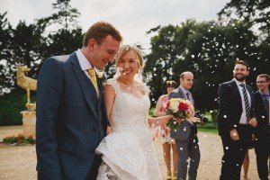 A joyful bride and groom, dressed in a white lace gown and a dark suit, smile as confetti flutters around them. The bride holds a colorful bouquet. Captured artfully by Somerset wedding photographers, friends in semi-formal attire celebrate against the charming garden backdrop.