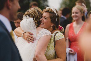A bride in a white dress and veil warmly hugs a woman in a yellow dress, both smiling amid the joyous gathering. Other well-dressed guests are seen chatting in the blurred outdoor setting, all perfectly captured by an expert Bristol wedding photographer.