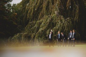 Five men in suits stand on a grassy field in front of tall, lush trees. One man raises his arm in mid-gesture. The scene has a serene, natural backdrop with the trees providing shade and depth, captured perfectly by Bristol wedding photographers with a documentary flair.