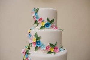 A three-tiered white cake is adorned with colorful flowers in shades of pink, blue, and yellow, interspersed with green leaves. The floral decorations cascade elegantly down the side, ready to be captured by Somerset wedding photographers amidst the celebration.