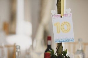 A table centerpiece at a Somerset wedding, with a small card hanging from a branch displaying the number 10 in yellow. The card features decorative designs and two heart-shaped clips, while bottles and a blurred dining setup linger in the background—perfect for documentary wedding photographers to capture.