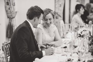 A couple sits at an elegantly decorated wedding reception table, beautifully captured by Somerset wedding photographers. The bride, in a white dress, smiles at the groom holding her hand. Glasses and plates gleam on the table, with guests blurred in the background.