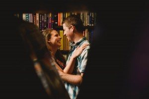 A couple standing close and smiling at each other in front of a bookshelf filled with colorful books, captured beautifully by Bristol wedding photographers. The scene is framed creatively with a blurred foreground.