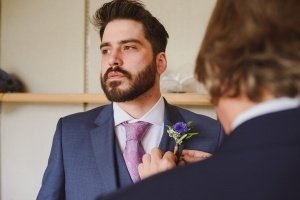 A man with a beard, dressed in a blue suit and pink tie, stands looking thoughtful as another person pins a boutonniere of purple flowers and greenery on his lapel. Captured by Bristol wedding photographers, the indoor setting features light-colored walls in the background.