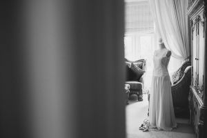 A black and white photo captures the essence of elegance, featuring a bridal dress on a mannequin in a serene room styled by Bristol wedding photographers. The lace-detailed gown graces the windows soft light, while shoes rest nearby and an ornate chair adds timeless charm to this documentary scene.