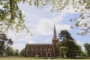 A historic church with arched windows and a tall spire is set in a lush green landscape, perfect for Bristol wedding photographers. Leaves from overhanging branches frame the scene, while scattered clouds dot the sky. Tall trees surround the church, enhancing this serene backdrop ideal for documentary wedding photographers.