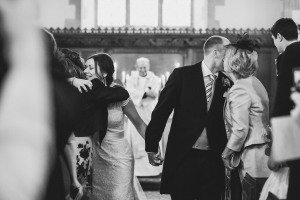 A bride and groom kiss while holding hands during a wedding ceremony, perfectly captured by a talented Cotswolds wedding photographer. The bride hugs a guest, while the groom receives a kiss from another guest. Candles and a smiling officiant add charm to the church setting.