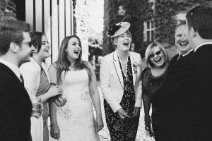 A black and white photo captures a wedding group laughing joyfully. The bride, radiant in her white dress, is surrounded by well-dressed guests near a vine-covered building. This candid moment showcases the skill of documentary wedding photographers in capturing Bristols celebratory spirit.