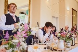 A man in a waistcoat and tie stands giving a speech at the banquet table, while another man sits with his hands covering his face, surrounded by guests. With colorful flowers and drinks, the scene is one wedding photographers in Bristol would love to capture for its candid emotion.