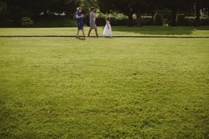 Three people stroll along a path in a large grassy area. The scene resembles a candid moment captured by Somerset wedding photographers. Two adults are on the left, one with a blue bag, while a child in a white dress walks on the right amid trees and bushes.