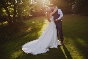 A bride and groom share a kiss on a sunlit lawn, captured brilliantly by Bristol wedding photographers. The bride dazzles in a long white gown, while the groom looks sharp in his dark suit. Sunlight filters through the trees, creating a warm glow and casting long shadows on the grass.