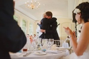 A groom embraces an older man, possibly his father, at a joyous wedding reception. Theyre surrounded by clapping guests. The elegantly decorated room features large windows and a chandelier. Tables are set with glassware and folded napkins, capturing the perfect scene for Bristol wedding photographers.