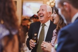 At the festive gathering, an older man in a suit and tie laughs heartily while holding a bottle. Surrounded by smiling guests engaged in lively conversation, you might think this joyful scene was captured by one of those talented documentary wedding photographers.