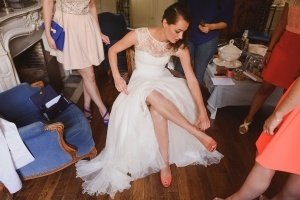 A bride in a lace bodice wedding dress adjusts her orange shoe, surrounded by friends in colorful outfits helping her prepare. The cozy room, captured beautifully by Bristol wedding photographers, features wooden floors and a small table in the background.