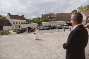 A bride in a white dress walks towards a man in a suit on a cobblestone plaza, an ideal moment for documentary wedding photographers. A row of parked cars lines the street against rustic buildings under a cloudy sky, as another person stands near the cars.