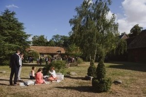 People gathered in a grassy outdoor area with trees and rustic buildings in the background, captured beautifully by Bristol wedding photographers. Some are seated on cushions on the ground, while others stand and converse under a clear blue sky. The setting appears relaxed and informal.
