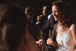 A bride in a white dress and lace top smiles while holding a champagne glass, captured perfectly by documentary wedding photographers. She is outdoors, surrounded by guests, as a woman in sunglasses laughs nearby. The background subtly blurs with people dressed in formal attire.