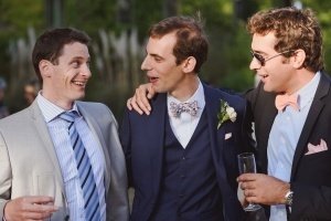 Three men in suits, likely captured by Somerset wedding photographers, are smiling and holding drinks outdoors. The man in the center sports a boutonniere and floral bow tie; his companions wear striped ties. They seem to be at a formal event, possibly a wedding.