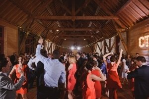 A lively wedding reception unfolds in a rustic wooden barn, captured perfectly by skilled documentary wedding photographers. Guests dance energetically in formal attire, with several women in red dresses. String lights and a large illuminated sign add warmth to the charming setting.