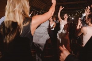 A bride in a white dress dances energetically on a crowded dance floor, captured perfectly by talented wedding photographers from Somerset. People around her are also dancing with arms raised, under string lights hanging from the ceiling, creating a festive atmosphere.