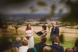 A bride in a white dress and a groom in a suit stand outside during a wedding ceremony as the officiant reads from a book. Wedding photographers from Bristol capture the moment, with guests seated and scenic fields and a charming town visible in the background.