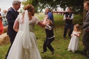 A bride in a white dress with a veil gives a high-five to a young boy jumping with excitement, while the groom stands nearby. A young girl in a white dress watches, as guests mingle in the background near trees and hay bales—a perfect moment captured by wedding photographers Somerset.