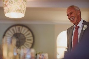 An older man in a grey suit with a white flower boutonniere smiles warmly in a softly lit room, captured expertly by wedding photographers Somerset. A large circular decorative piece hangs on the wall behind him, and a patterned lampshade is visible above. Glasses are blurred in the foreground.