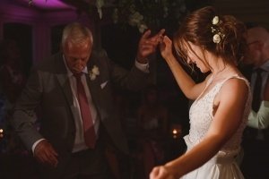 At a warmly lit event, an elderly man in a suit dances with a young woman in a white dress, her hair adorned with flowers. Both smile as Somerset wedding photographers capture this cherished moment amidst the dimly lit background filled with other guests.
