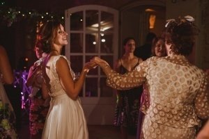 A bride in a white dress dances joyfully with a woman in a patterned outfit at a warmly lit indoor event captured by skilled Bristol wedding photographers. Other guests, dressed in vibrant attire, are visible amid string lights and a blurred doorway enhancing the festive atmosphere.