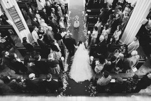 A black-and-white image captures a bride and groom walking down the church aisle, taken by skilled Bristol wedding photographers. Guests are seated on either side, while a child flower girl scatters petals ahead of them. The scene is beautifully viewed from above.