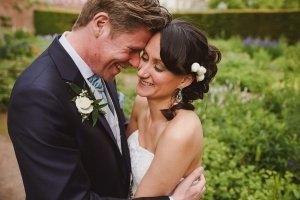 A couple in wedding attire embrace joyfully outdoors, captured beautifully by Bristol wedding photographers. The groom wears a suit with a white rose boutonniere, and the bride has flowers in her hair. They smile and touch foreheads in a charming garden setting.