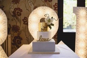A three-tiered white cake displays a gold top tier and a decorative white flower with greenery. Love is scripted in gold on the middle tier, perfect for capturing by Somerset wedding photographers. Large illuminated numbers form the backdrop, set against floral wallpaper.