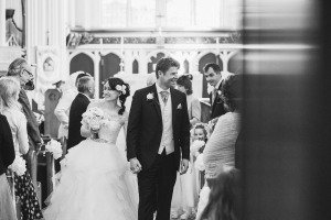 A bride and groom smile as they walk hand in hand down the aisle of a church, surrounded by smiling guests. Captured by Somerset wedding photographers, the bride holds a bouquet of flowers, and the groom wears a suit and tie. The scene is joyful and filled with celebration.