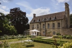 A large, rustic stone house with a steep, tiled roof is surrounded by a lush garden, perfect for a wedding photoshoot. A white parasol shades a table set with chairs on the lawn. The pathway leads to the entrance, and tall trees stand in the background under a partly cloudy sky.