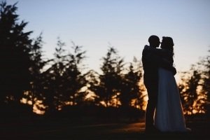A couple is silhouetted against a vibrant sunset, embracing lovingly with trees in the background. The sky is a gradient of deep blue to warm orange, creating a romantic and serene atmosphere—a perfect moment captured by documentary wedding photographers in the Cotswolds.