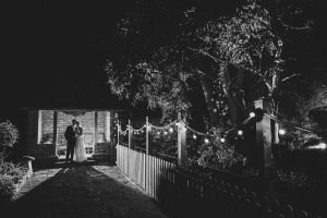 A couple stands embraced under a canopy of string lights, their romantic nighttime moment captured in black and white by Bristol wedding photographers. The path leads to a quaint, rustic building framed by trees, enhancing the serene and intimate atmosphere.