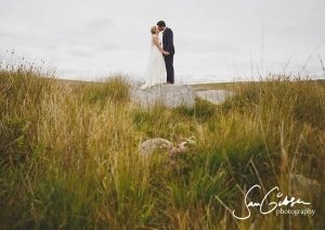 A bride and groom stand on a large rock in a field, surrounded by tall grass and wildflowers, captured beautifully by Somerset wedding photographers. They hold hands and kiss under a cloudy sky, with the photographers logo visible in the bottom right corner.