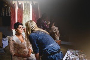A makeup artist applies eyeshadow to a seated woman in a floral robe, set in a dimly lit room. Like wedding photographers in Bristol, capturing candid moments, another woman is being styled by the window among hanging garments. A table brims with makeup supplies in the foreground.