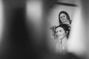 Black and white image of a woman sitting as another styles her hair with a comb, captured through a soft-focus foreground. This candid and intimate scene showcases the artistry of wedding photographer Cotswolds, specializing in capturing timeless moments.