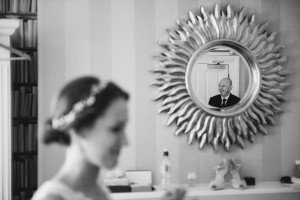 A bride is in the foreground, slightly out of focus, brilliantly captured by a wedding photographer from the Cotswolds. In the sun-shaped mirror on the wall behind her, a man in a suit appears. The room features striped wallpaper and shelves lined with books.