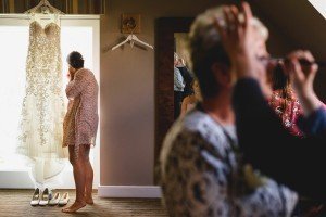 A bride stands in a floral robe, gazing out the window with her lace wedding dress and white shoes below. In the foreground, as Bristol wedding photographers capture candid moments, someone is having makeup applied. A large mirror adorns the wall, reflecting the days elegance and anticipation.