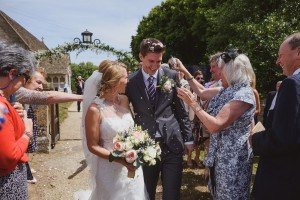 A bride and groom walk joyfully outdoors, surrounded by smiling guests throwing confetti, as a Somerset wedding photographer captures the moment. The bride holds a bouquet of pink and white flowers, and the groom wears a dark suit with a striped tie. Greenery and a quaint building are in the background.