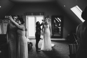 A bride in a fitted lace gown stands smiling as her hair is adjusted, captured beautifully by a wedding photographer from Cotswolds. Two bridesmaids in elegant dresses take a selfie on the left, while another stands to the right. The room is softly lit by a window and features a large mirror.