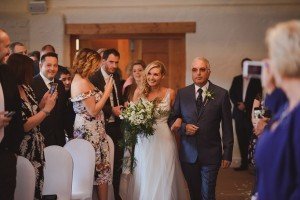 A bride in a white dress holding a bouquet walks down the aisle with an older man in a suit. Guests, with smiles all around, take photos. The indoor ceremony, beautifully captured by talented Somerset wedding photographers, features white chairs and wooden doors.