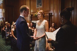 A bride and groom stand facing each other, holding hands during their wedding ceremony. A person officiates the ceremony while reading from a book. Guests are seated in the wooden-paneled room, watching the couple, as Bristol wedding photographers capture every heartfelt moment.