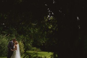 A bride and groom stand closely together in an intimate pose, surrounded by lush greenery. Captured by Somerset wedding photographers, the bride is wearing a white gown with a veil, and the groom is in a suit. The scene is serene, with soft, natural lighting filtering through the trees.