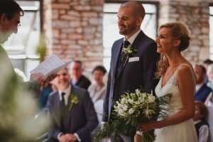 A bride and groom stand together indoors at a wedding ceremony, facing an officiant holding a paper. The bride clutches a bouquet of white flowers, smiling joyfully. Expertly captured by documentary wedding photographers, guests are seated against a rustic brick wall in the background.