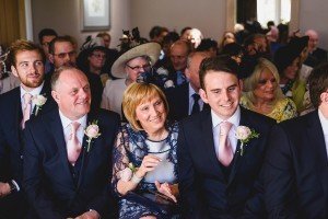 A group of people, smartly dressed, sit closely together in what appears to be a wedding ceremony. They are wearing suits, ties, and floral corsages. A woman in the center wears a blue lace dress and hat. As captured by wedding photographers Somerset, they look towards the altar, smiling.